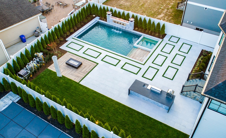 Aerial view of a modern backyard with a rectangular pool, hot tub, lounge area, grill station, and geometric grass and stone design, all enclosed by a white fence and tall shrubs.