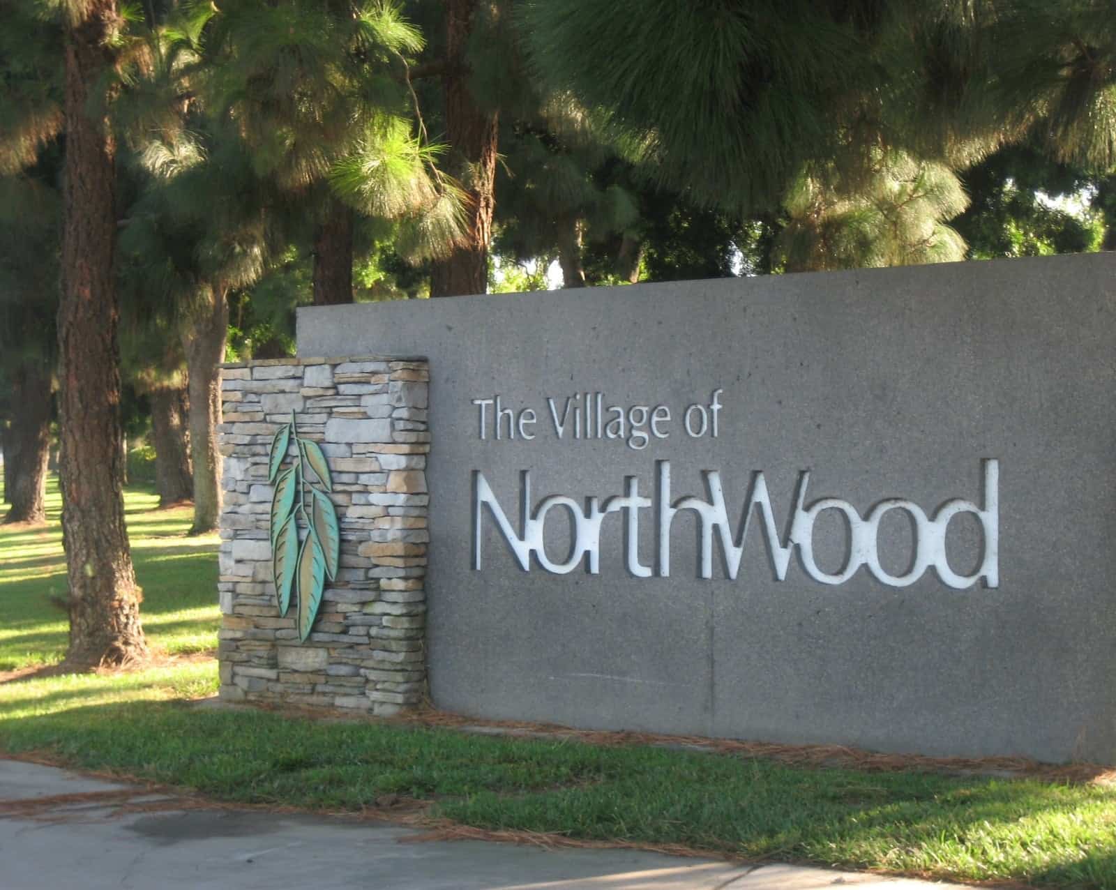 A low stone wall bearing the words The Village of Northwood in raised gray letters, framed by shrubs and tall eucalyptus trees in Irvine.