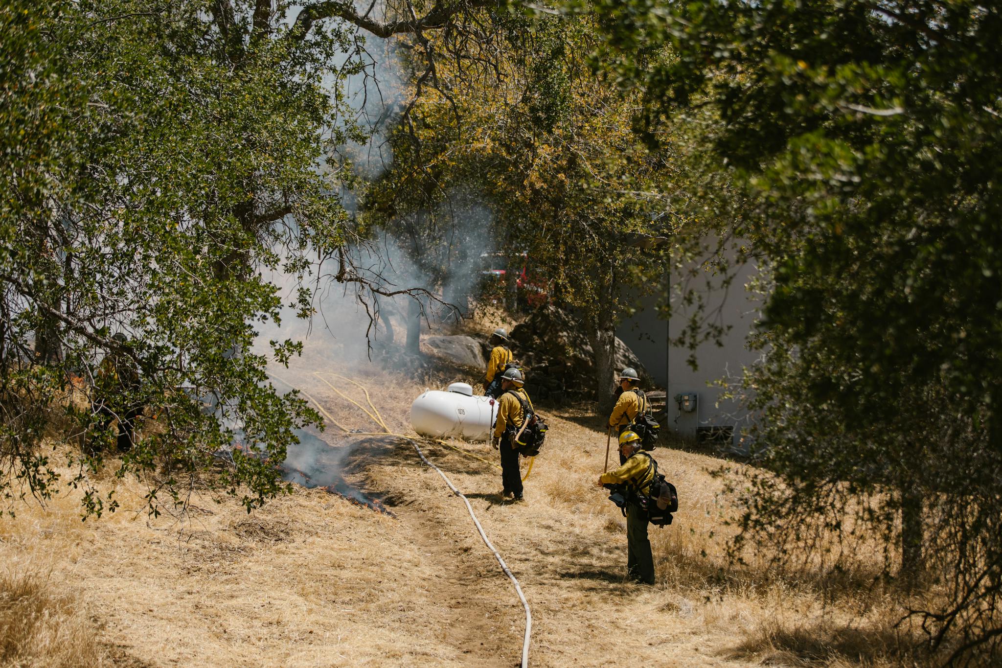 Firefighters in protective gear combat a wildfire in a dry, wooded area with visible smoke.