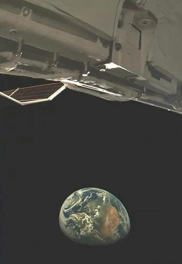 Earth as seen from space, with a view of the X-37B's exterior. The blue and brown continents of Earth are visible against the black expanse, with a portion of the orbital vehicle in the foreground. This is a Space Force mission.