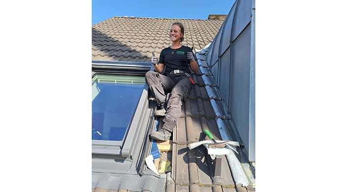 Eine Frau sitzt lächelnd auf einem Dach und zeigt mit beiden Händen Daumen hoch. Sie trägt Arbeitskleidung und Handschuhe, Werkzeug ist sichtbar.