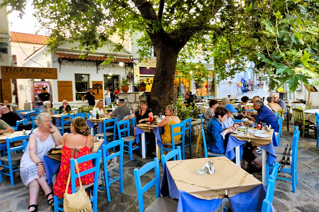Viele Menschen sitzen in Skopelos auf blauen Stühlen einer Taverne rund um einen großen Baum