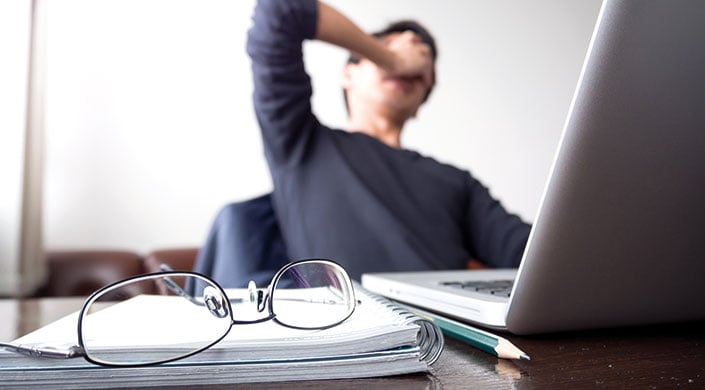 Brille auf einem Notizbuch auf einem Schreibtisch, im Hintergrund eine Person, die müde wirkt und sich das Gesicht reibt.