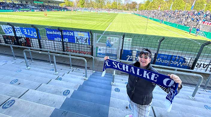 Blick von einer Stadiontribüne auf ein Fußballfeld, mit einem blauen Fan-Schal, einem Zaun sowie Werbebannern im Vordergrund.