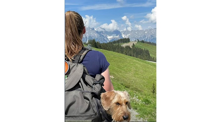 Person mit Rucksack und kleinem Hund auf einer grünen Wiese, im Hintergrund Berge und blauer Himmel mit Wolken.