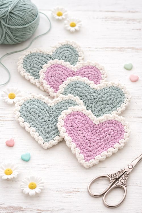 Pastel pink and teal crocheted heart coasters with white borders on a white wood surface, surrounded by yarn, daisies, scissors, and candies. Free crochet pattern