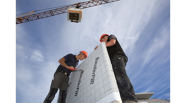 Zwei Bauarbeiter mit orangefarbenen Helmen halten eine große Dämmplatte mit der Aufschrift "BauderPIR" auf einer Baustelle, Kran im Hintergrund.
