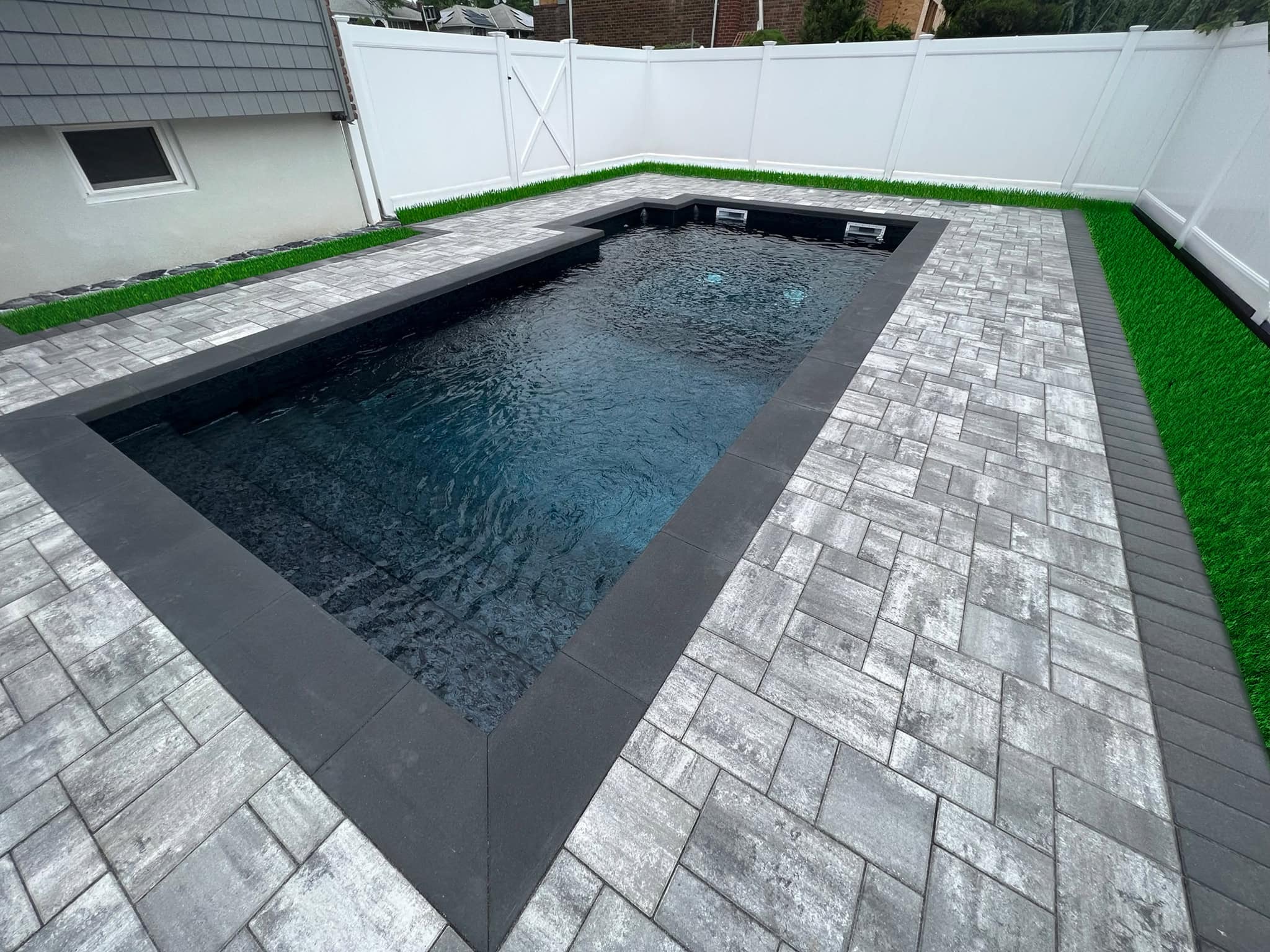 Rectangular backyard pool surrounded by gray stone pavers and artificial grass, enclosed by a white privacy fence.