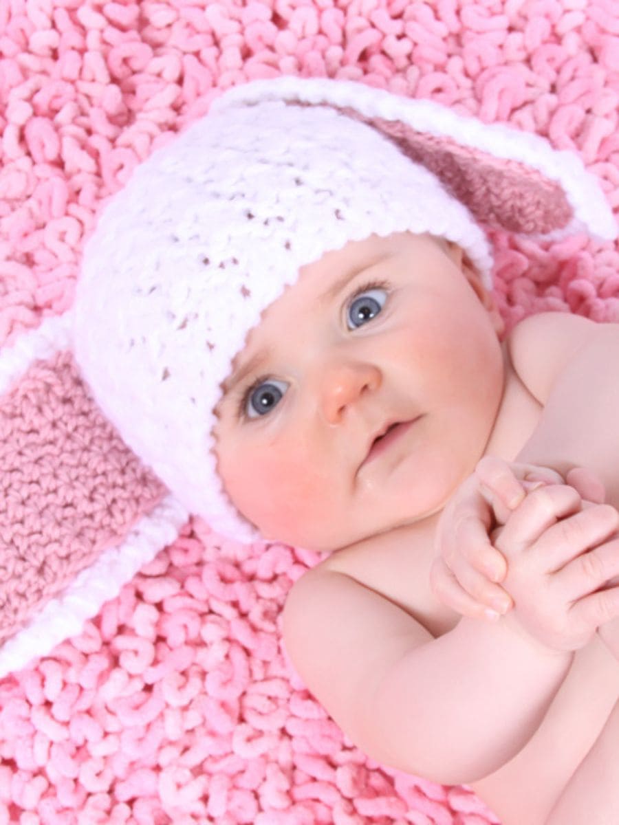 baby wearing white crochet bunny hat with large floppy ears laying on a pink carpet