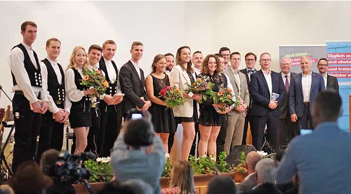 Mehrere Personen in festlicher Kleidung stehen nebeneinander auf einer Bühne. Einige halten Blumensträuße. Im Vordergrund fotografieren Zuschauer die Szene.