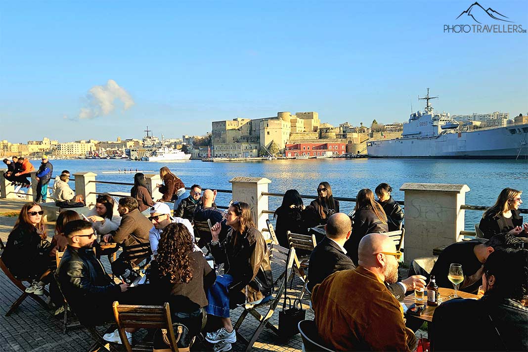 Viele Menschen sitzen in der Sonne in einem Cafe mit Blick auf die Altstadt von Brindisi