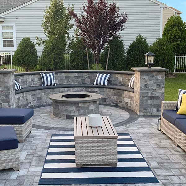 Outdoor patio featuring a circular stone fire pit, surrounded by benches with striped cushions, coordinate in a set with a square coffee table and blue-and-white area rug.