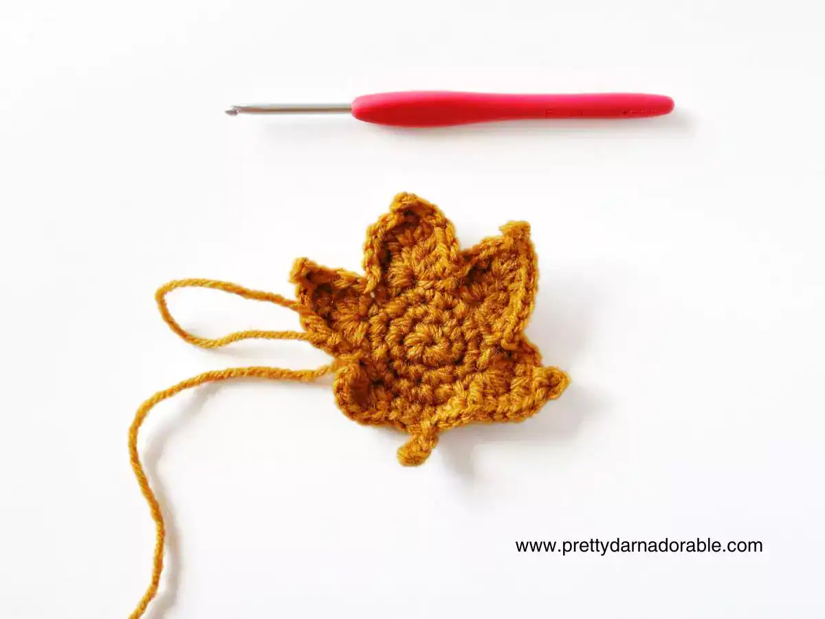Yellow crocheted maple leaf with loose yarn ends on white surface beside red crochet hook; free pattern at prettydarnadorable.com. Free crochet pattern