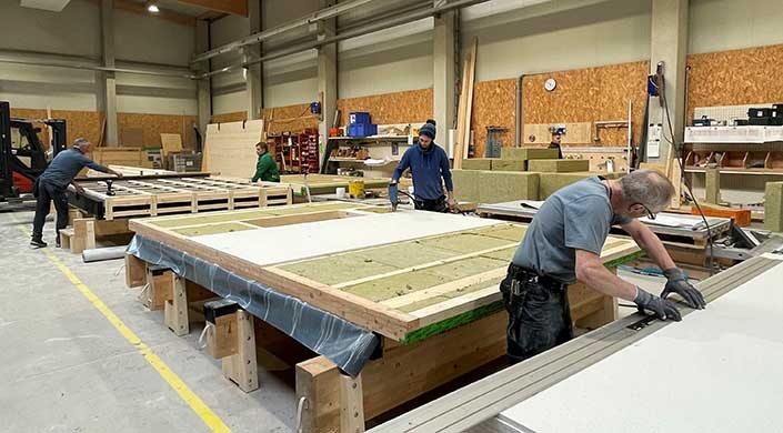 Ein Mann arbeitet in einer Werkstatt an einer Holzkonstruktion mit Dämmmaterial, umgeben von Werkzeugen und Baumaterialien.