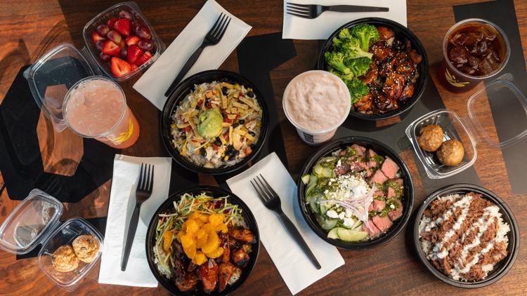 Overhead spread of multiple Konala protein bowls, snacks, fresh fruit, and beverages on a wooden table