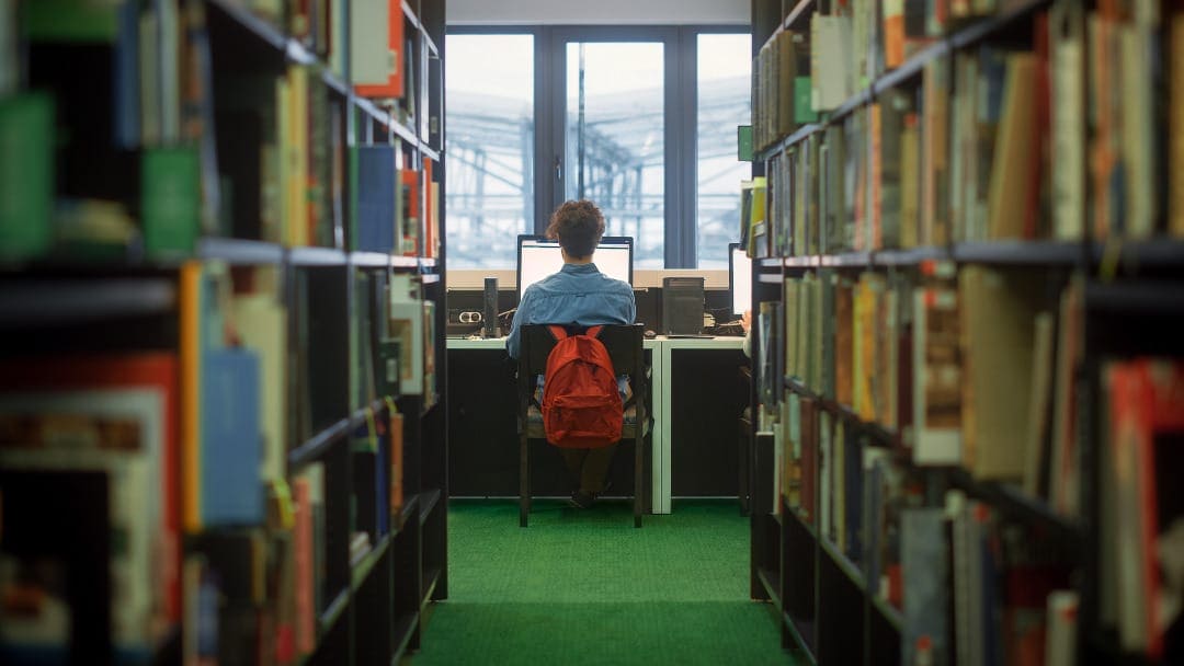 Rezension schreiben Promotion Rezension schreiben Promotion
Das Bild zeigt eine Person, die zwischen hohen Regalen voller Bücher in einer Bibliothek sitzt. Die Person ist von hinten zu sehen, sitzt an einem Schreibtisch und ist auf einen Computerbildschirm konzentriert. Sie trägt ein blaues Hemd und hat einen roten Rucksack, der auf dem Boden neben dem Stuhl steht. Das natürliche Licht scheint durch ein großes Fenster im Hintergrund herein, das einen Ausblick auf eine Struktur außerhalb des Gebäudes bietet. Die Umgebung wirkt ruhig und still, typisch für eine Bibliothek, wo Konzentration und Lernen gefördert werden. Der grüne Teppich auf dem Boden liefert einen angenehmen Kontrast zu den bunten Buchrücken und der insgesamt ruhigen Farbpalette des Raumes.