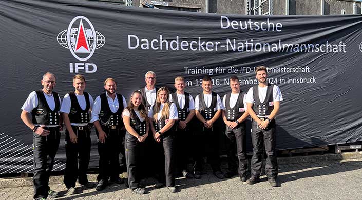 Fünf Personen in traditioneller deutscher Handwerkskleidung stehen vor einem schwarzen Banner mit weißer Aufschrift und lächeln in die Kamera.