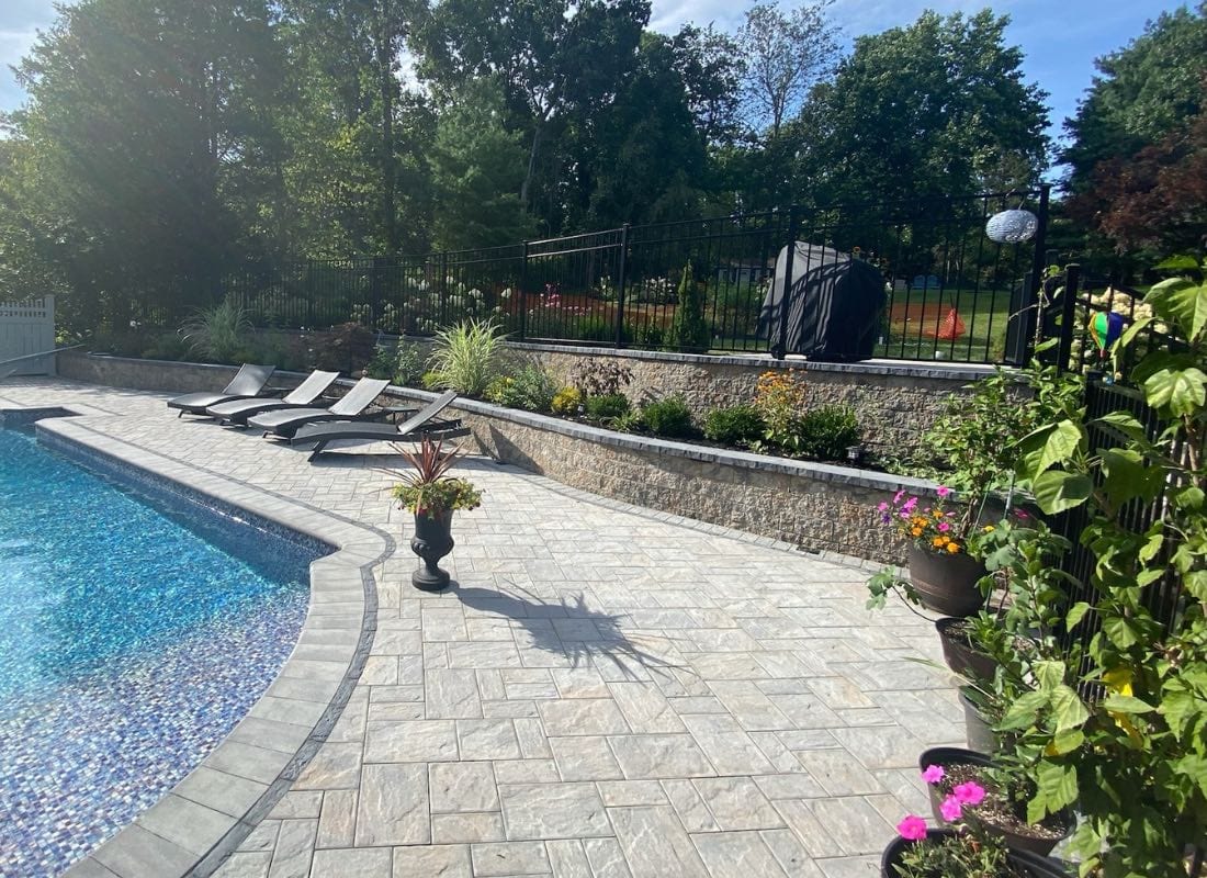 A sunny backyard with a curved swimming pool, stone patio, sun loungers, and a view of a court behind a metal fence.