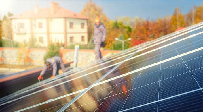 Solarmodule im Vordergrund, unscharfe Person mit Schutzhelm im Hintergrund, Gebäude und herbstliche Bäume sichtbar.