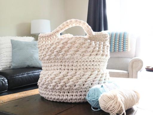 A thick, hand-crafted white basket rests on a wooden table beside blue and cream yarn balls, with a couch in the background featuring pillows and a crocheted blanket draped over the armrest. Free crochet pattern