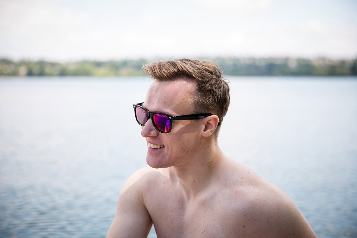 A person with sunglasses smiles while standing shirtless by a body of water, with a blurred shoreline in the background.