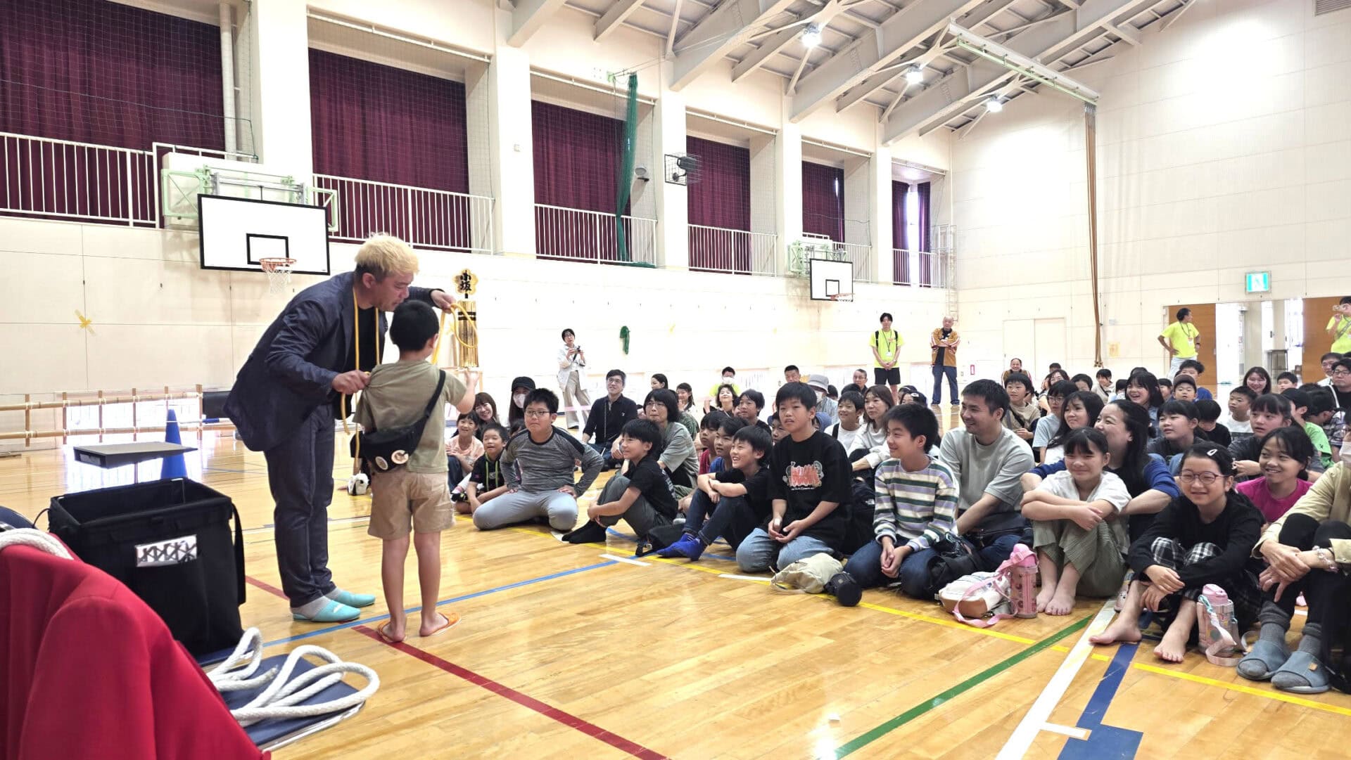 金沢市小坂小学校で2年連続マジックショー!昨年との違いに感じた「文化を育てる」ということ 5 マジシャン・コンプレッサーが観客席の男の子と至近距離で向き合い、マジックを行っている。周りの観客は笑顔で見守っている。