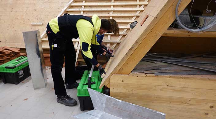 Eine Person in Arbeitskleidung und Handschuhen arbeitet an einer Holzkonstruktion, wahrscheinlich auf einer Baustelle im Innenbereich.