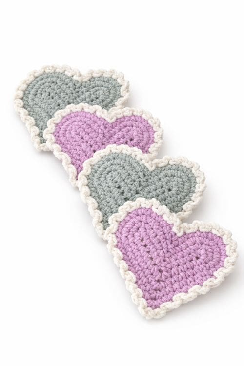 Four heart-shaped crochet coasters in light purple and gray with white scalloped edges, arranged in a slightly overlapping row. Free crochet pattern