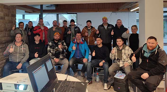 Eine Gruppe von Menschen sitzt und steht zusammen drinnen und zeigt Daumen hoch, einige lächeln in die Kamera.