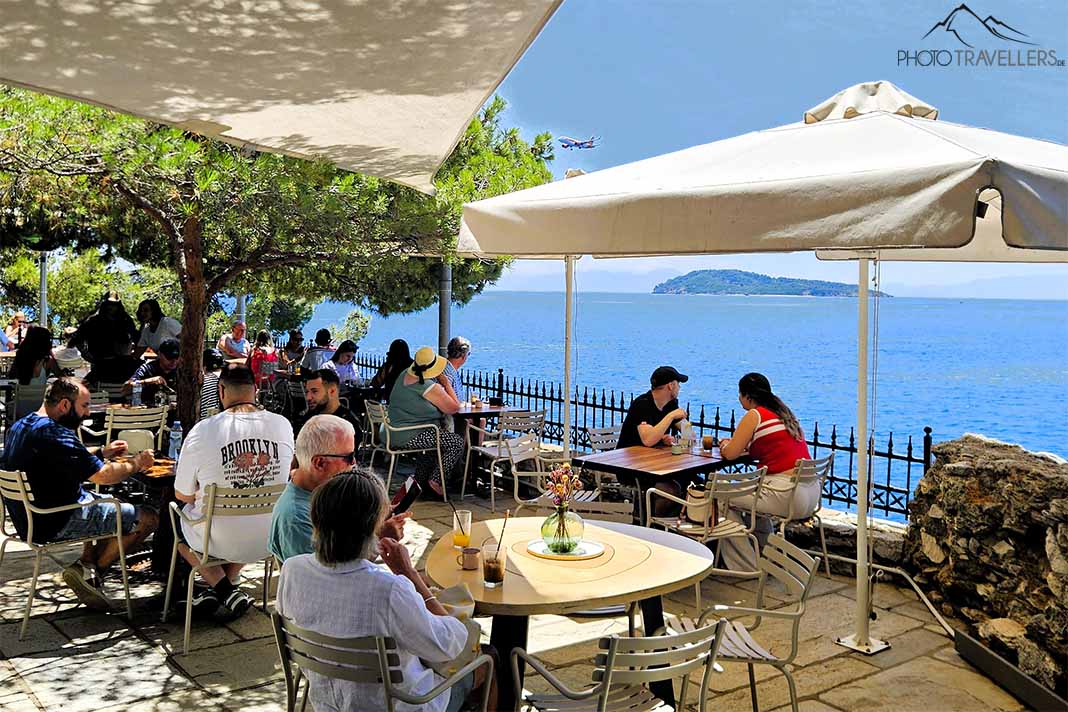 Menschen sitzen unter Sonnenschirmen und Bäumen im Bourtzi Café über dem Meer