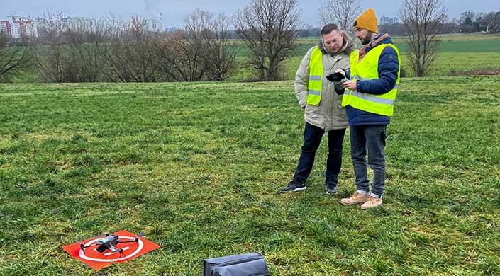 Person steht auf einer Wiese im Freien, mit kahlen Bäumen im Hintergrund und einer Tasche auf dem Gras.