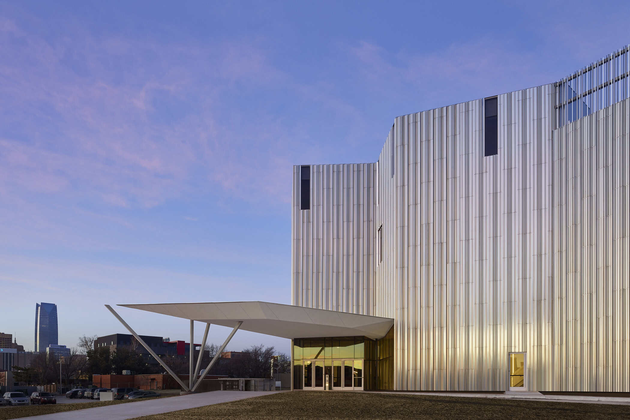 Exterior view of the Oklahoma Contemporary Arts Center, featuring the aluminum-clad façade and cantilevered canopy at sunrise, with the Oklahoma City skyline in the background.
