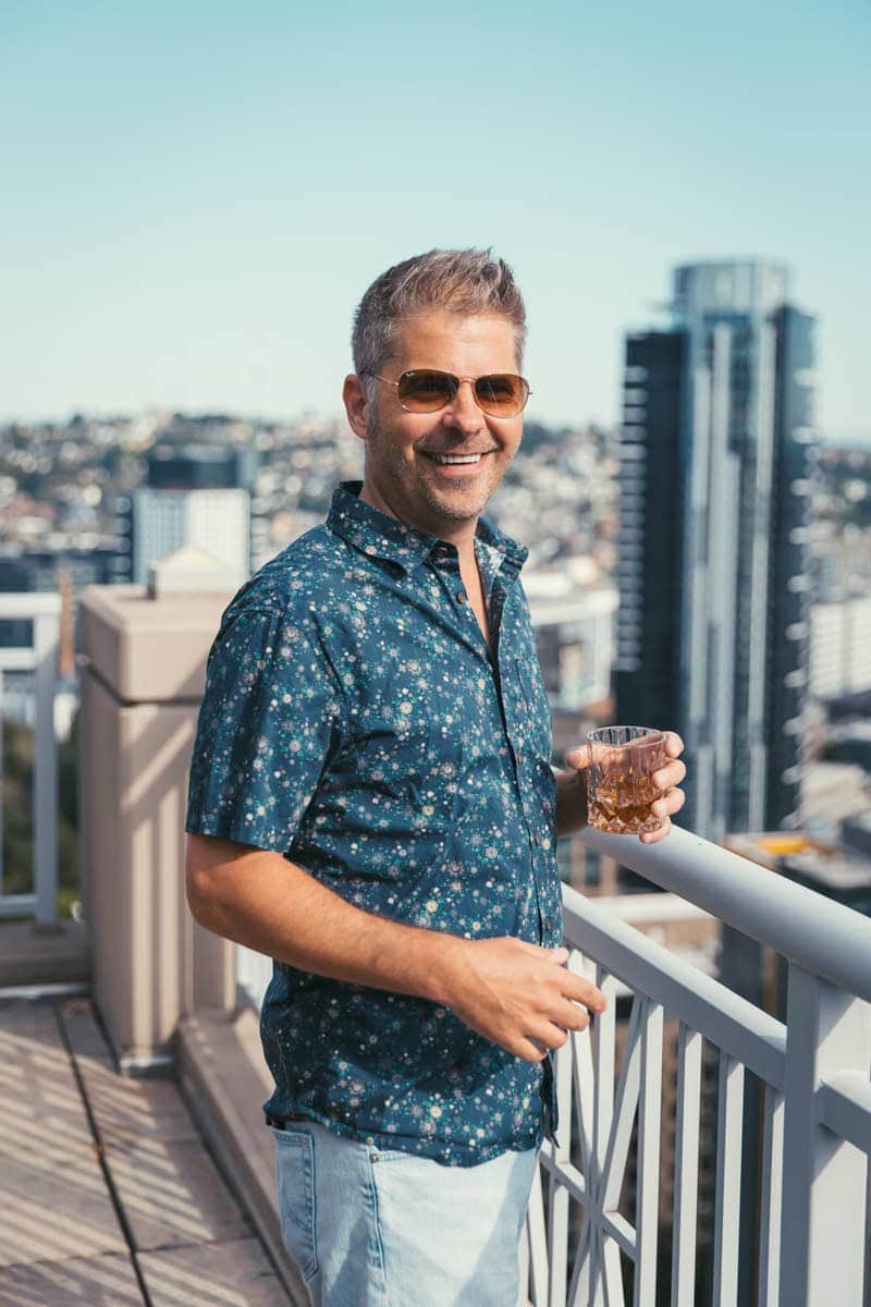 A man in sunglasses and a floral shirt stands on a rooftop balcony, holding a drink, with a cityscape and tall buildings in the background.