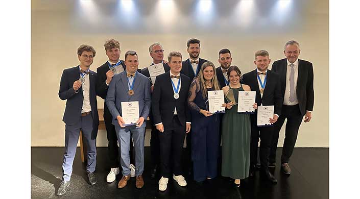 Eine Gruppe von Menschen in formeller Kleidung hält Urkunden und Medaillen und posiert für ein Foto bei einer Preisverleihung oder einer offiziellen Veranstaltung.