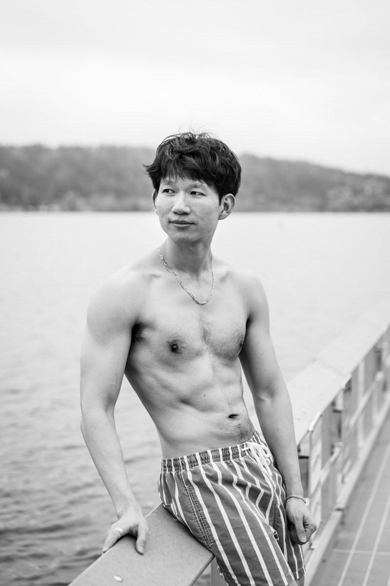 A person with short hair stands shirtless by a railing near a body of water, wearing striped shorts.