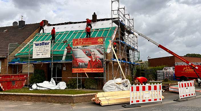 Arbeiter decken ein Hausdach mit Folie ab, ein Werbebanner mit einem Mann in roter Kleidung hängt am Baugerüst, Bauholz liegt vorne.