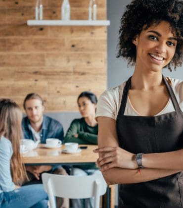 Smiling woman in apron stands in foreground of café, with three people seated at a table in the background, drinking coffee.
