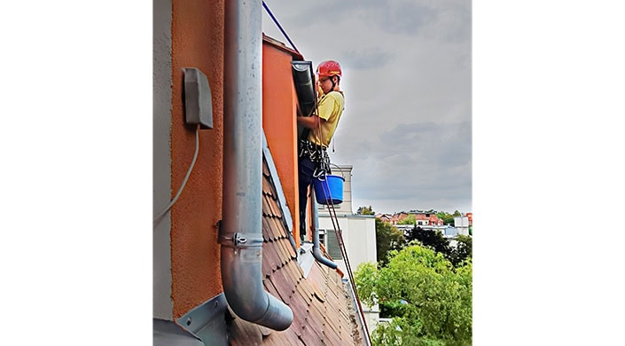 Ein Arbeiter mit Helm reinigt eine Regenrinne an einem Hausdach, gesichert mit Seilen und Ausrüstung, bei bewölktem Himmel.