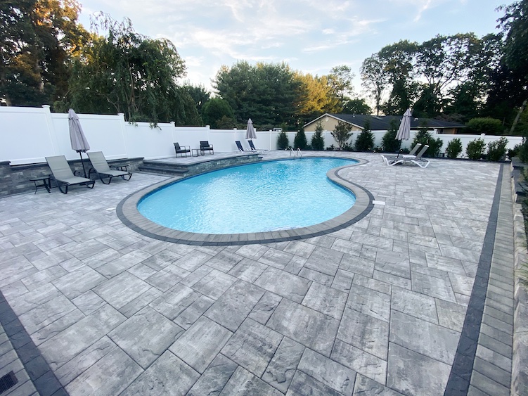 An outdoor swimming pool with curved edges is surrounded by gray stone pavers, lounge chairs, and umbrellas, with trees and a white fence in the background.