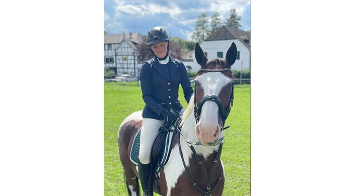 Eine Frau in Reitkleidung sitzt auf einem braun-weißen Pferd im Freien. Sie trägt einen Helm und lächelt, im Hintergrund ist ein Gebäude zu sehen.