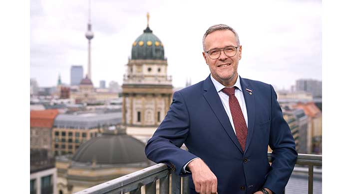 Ein Mann im blauen Anzug steht auf einem Balkon mit historischer Kuppel im Hintergrund.