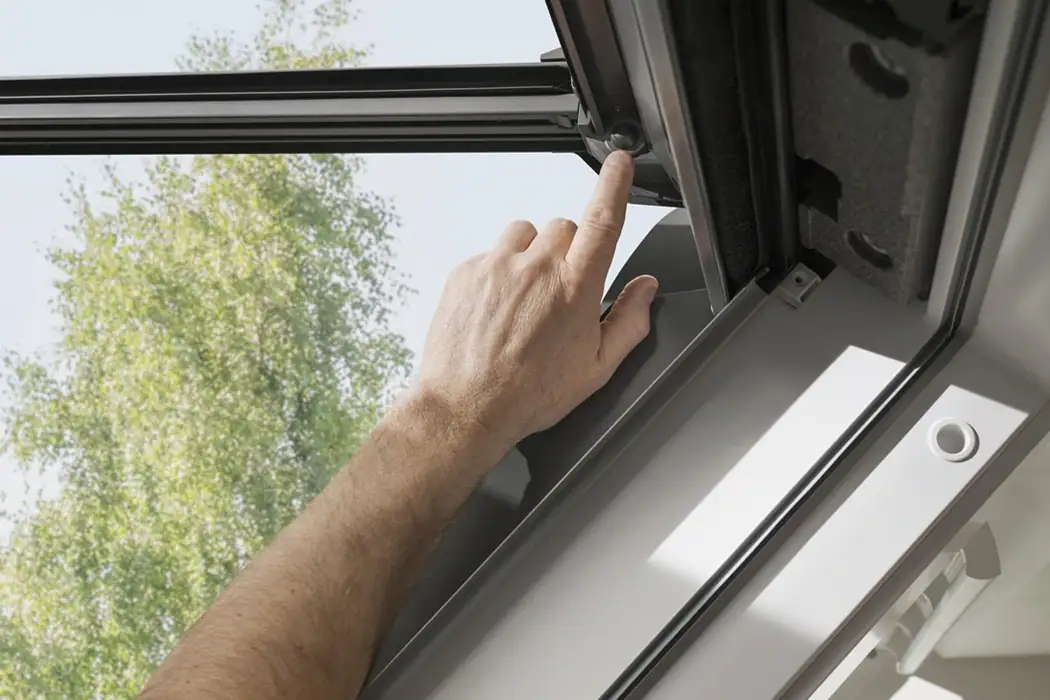 Eine Hand drückt auf einen Knopf am Fensterrahmen eines geöffneten Dachfensters, im Hintergrund ist ein grüner Baum zu sehen.
