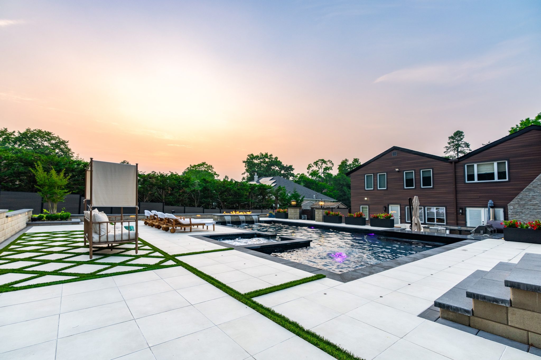 Modern backyard with a large pool, lounge chairs, geometric tile patio, and contemporary house at sunset.