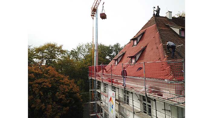Ein Baukran hebt Material auf ein Gerüst neben einem teils renovierten Gebäude mit rotem Dach. Bäume sind im Hintergrund sichtbar.