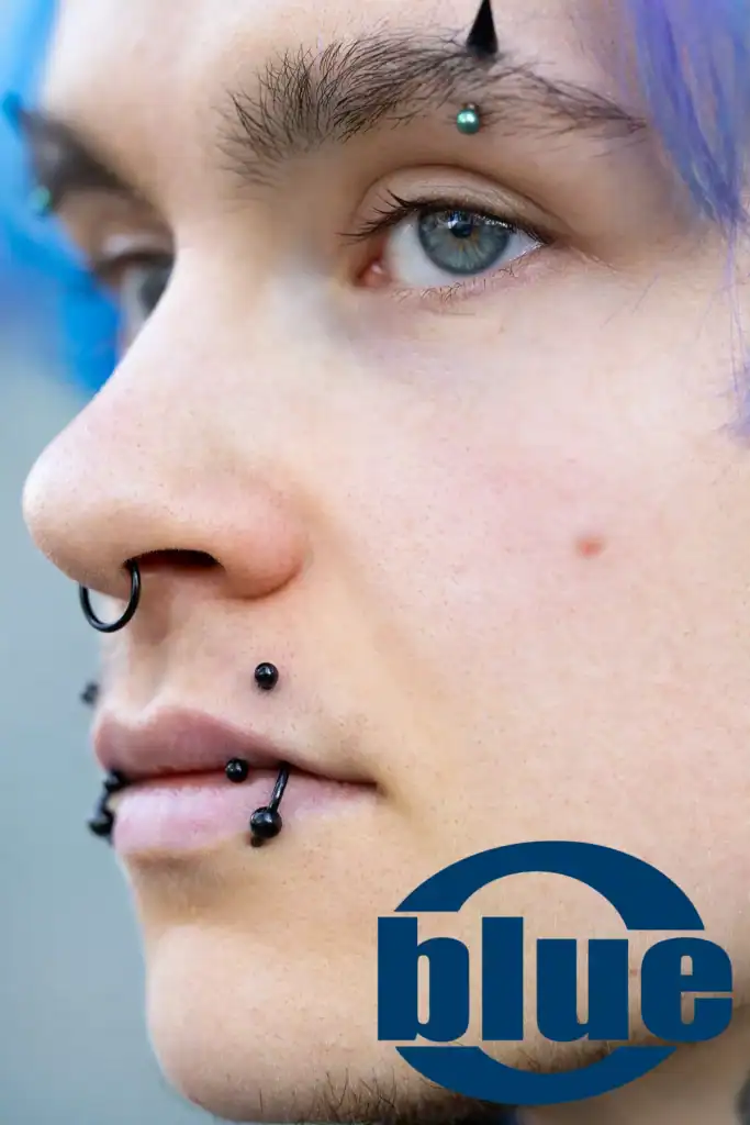 Close-up of a person with blue hair and multiple facial piercings (nostril, lip) and piercing jewelry, with a blue logo partially visible on the chin area.