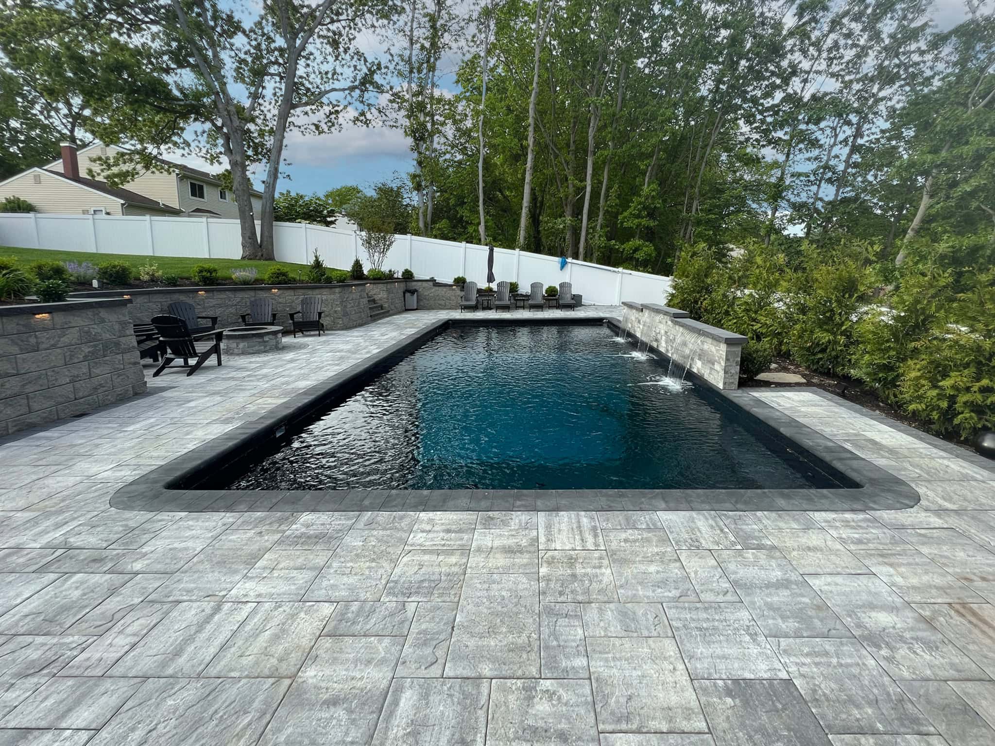 Rectangular in-ground pool with dark water, surrounded by gray stone tiles, black lounge chairs, and landscaped greenery under a partly cloudy sky.