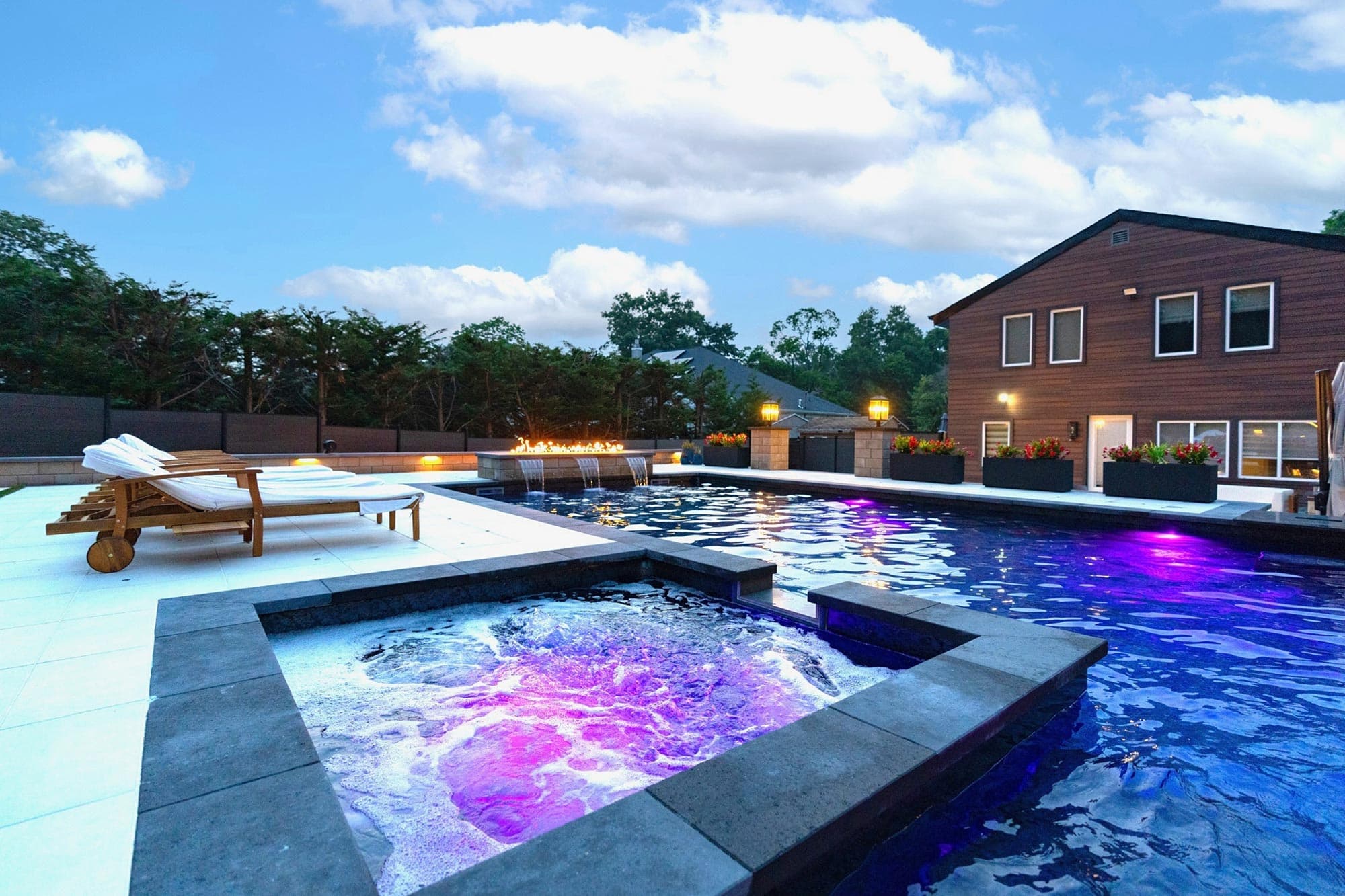 Modern backyard with a jacuzzi and swimming pool, poolside lounge chairs, and a contemporary house in the background under a partly cloudy sky at dusk.