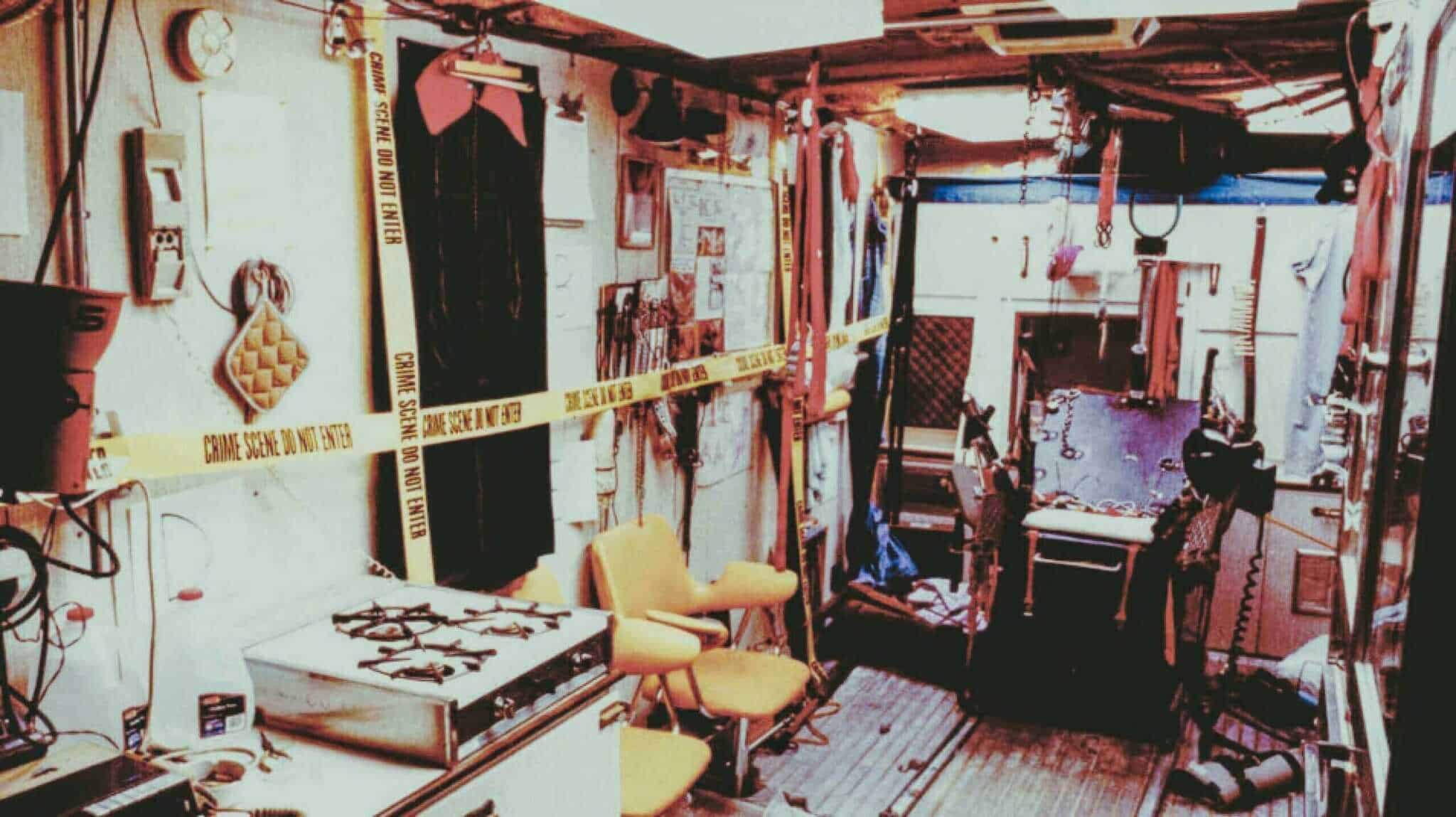 Interior of a cluttered trailer room strung with crime scene tape, tools and a chair.