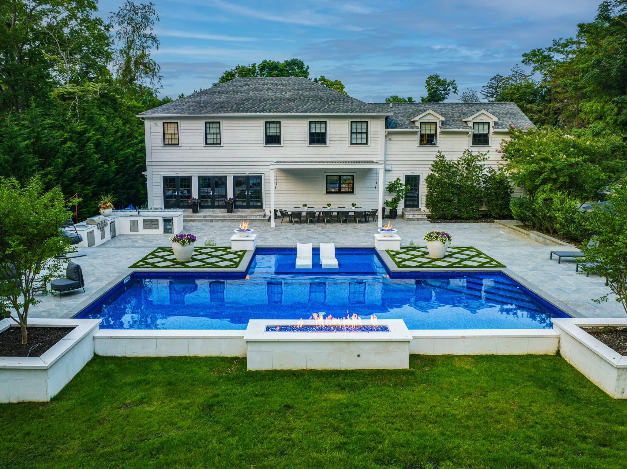 A modern backyard features a rectangular pool with water features, fire pits, lounge chairs, and a large two-story house in the background surrounded by trees.
