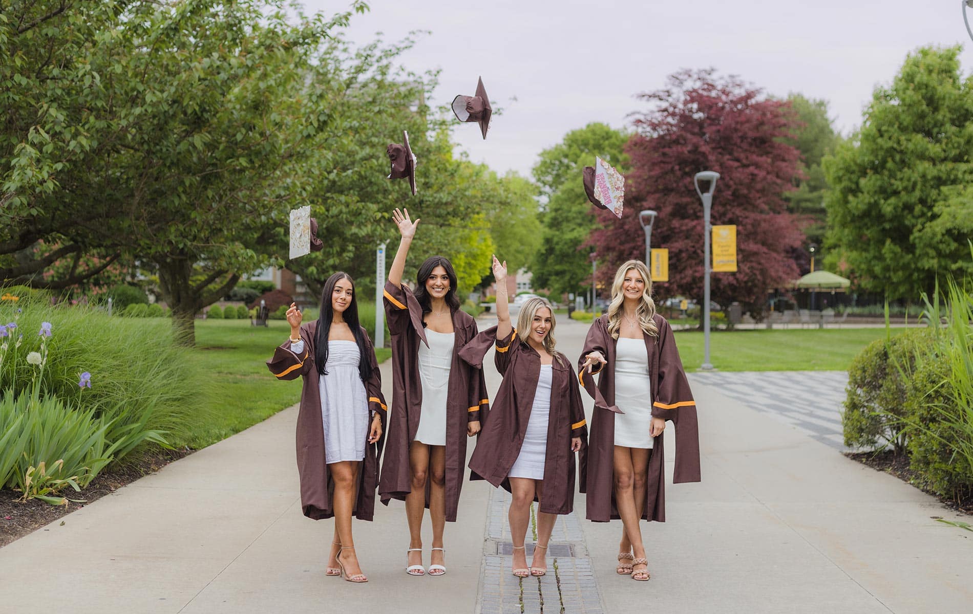 Four women in graduation gowns and white dresses stand outdoors on a pathway, smiling and tossing their caps into the air. Captured by a Long Island photographer, trees and greenery provide a beautiful backdrop to this joyful moment.